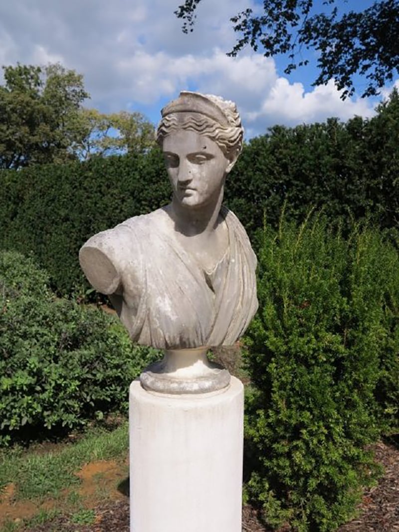 This photograph shows a grey marble ¾ bust of a woman with her face turned in a profile. The bust has drapery over her chest, and no arms past the shoulder. She has wavy, pulled back hair and a tiara. There is greenery and a blue sky with clouds in the background.