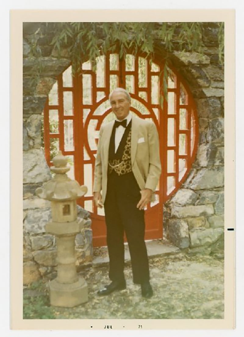 A color photograph of Julian Wood Glass Jr. standing in front of a red, round, wooden, gate surrounded by a stone wall. Light shows through the rectangular shapes that make up the gate. Green bamboo hangs down in the top of the image. Julian wears a tan suit jacket, black pants, a black bowtie, and a black and brown spotted vest. He faces the camera smiling.